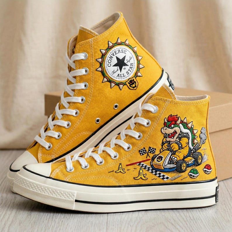 Puede incluir: Zapatillas altas Converse Chuck Taylor All Star de color amarillo mostaza. Un zapato presenta un personaje de Bowser conduciendo un kart, con banderas a cuadros y pieles de pl&aacute;tano. El otro zapato tiene un logotipo de Converse con el texto "All Star".