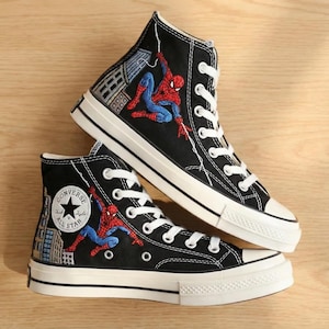 Puede incluir: Zapatillas altas negras con cordones y suelas blancas. Las zapatillas presentan un diseño bordado de Spider-Man con un fondo de paisaje urbano. El logotipo de Converse All Star es visible en el lateral.