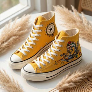 Puede incluir: Un par de zapatillas Converse All Star altas amarillas con cordones blancos y puntera blanca. Un zapato presenta un gr&aacute;fico de Bowser. El logotipo de Converse est&aacute; en el lateral. Los zapatos est&aacute;n sobre una superficie blanca.