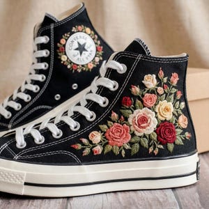 May include: Black high-top sneakers with white laces and soles. The sneakers feature embroidered floral designs in shades of red, pink, and yellow. One shoe has a Converse logo with floral accents.