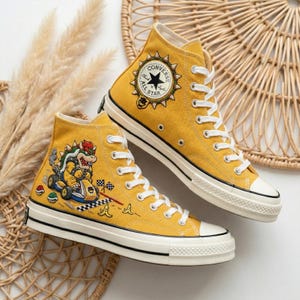 Puede incluir: Zapatillas altas de color amarillo mostaza con cordones y suelas blancas. Una zapatilla presenta un gr&aacute;fico de Bowser conduciendo un kart, la otra tiene un parche de Converse All Star. Las zapatillas est&aacute;n decoradas con un tema de videojuego.