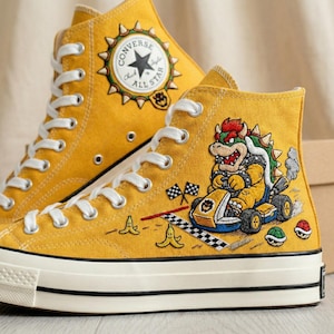 Puede incluir: Zapatillas altas de color amarillo mostaza con cordones y suelas blancas. Las zapatillas presentan un dise&ntilde;o del personaje Bowser en el lateral, con detalles de banderas a cuadros y c&aacute;scaras de pl&aacute;tano. El logo de Converse All Star est&aacute; en el tobillo.