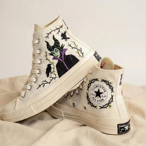 Puede incluir: Zapatillas altas color crema con diseños bordados. Un zapato presenta un retrato de personaje, el otro una corona con el logo de Converse All Star. Los zapatos tienen cordones blancos y suela de goma.