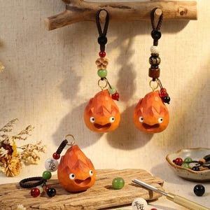 Può includere: Portachiavi in legno arancione a forma di demone del fuoco dei cartoni animati, con dettagli intagliati e facce sorridenti. Ogni portachiavi ha un filo di perline decorativo con un cordino nero. Un portachiavi poggia su una tavola di legno.