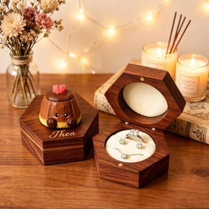May include: Two hexagonal dark wood jewellery boxes with a smooth finish. One box is closed and topped with a small wooden character figurine with a red heart, featuring the name Thea engraved. The other box is open, revealing white velvet lining.