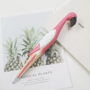 Puede incluir: Un bol&iacute;grafo con forma de flamenco rosa, con pico negro y detalles blancos. El bol&iacute;grafo es de madera y descansa sobre un papel con el texto "TROPICAL PLANTS". Un instrumento de escritura &uacute;nico.