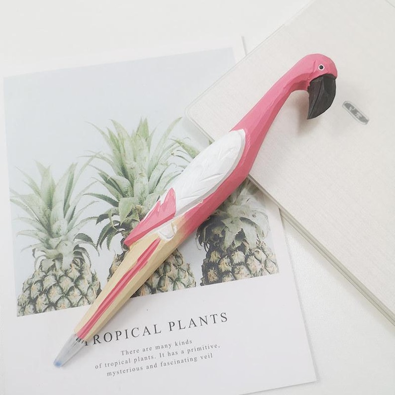 Puede incluir: Un bol&iacute;grafo con forma de flamenco rosa, con pico negro y detalles blancos. El bol&iacute;grafo es de madera y descansa sobre un papel con el texto "TROPICAL PLANTS". Un instrumento de escritura &uacute;nico.