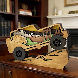 Può includere: Una scultura in legno a strati di un veicolo 4x4 che scala un terreno roccioso. L'interno presenta una scena di montagna e foresta con alberi, montagne e un cervo. Le ruote sono nere e il legno ha una finitura naturale.