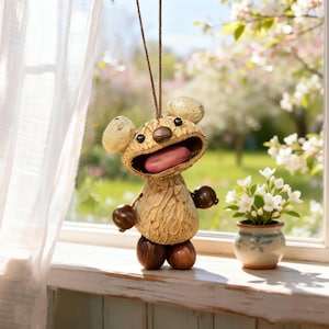 May include: A whimsical, handcrafted wooden bear ornament with a wide open mouth and a pink tongue. The bear has large, round ears and small, round arms and feet. It hangs from a brown string. A small vase of white flowers sits nearby.