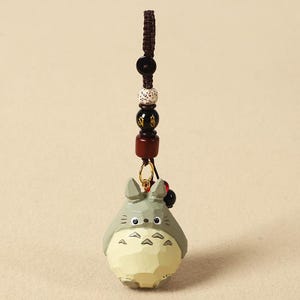 May include: A wooden Totoro keychain with a brown cord and decorative beads. The Totoro figure is gray with a cream-colored belly and black eyes. The keychain has a gold-colored ring.