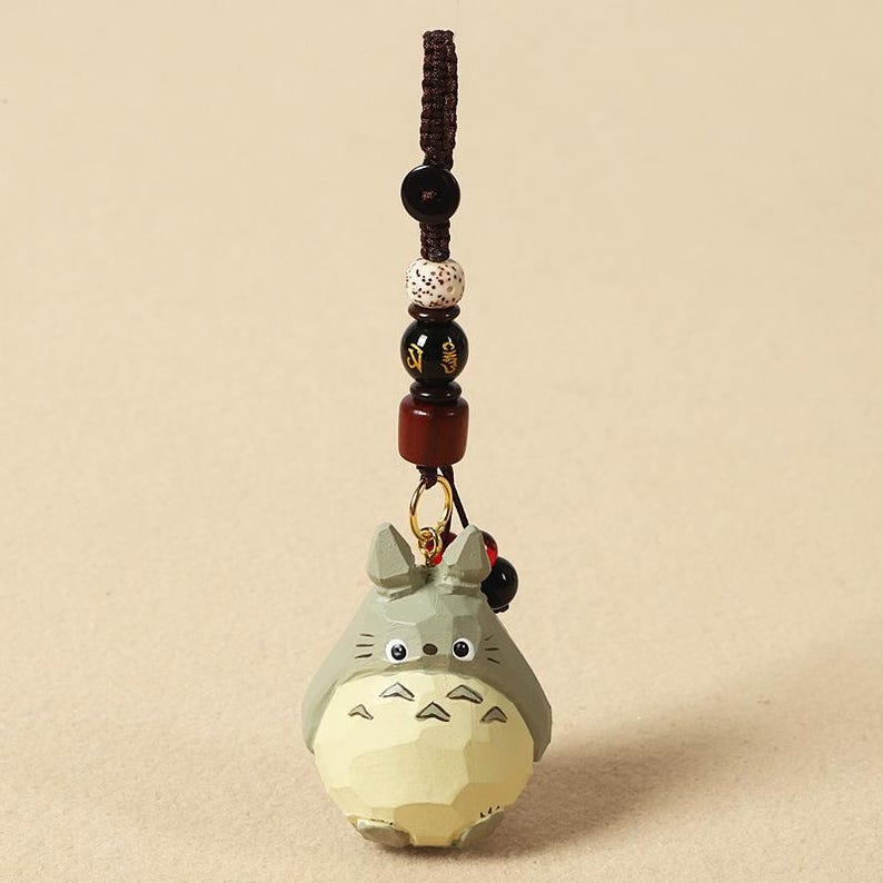 May include: A wooden Totoro keychain with a brown cord and decorative beads. The Totoro figure is gray with a cream-colored belly and black eyes. The keychain has a gold-colored ring.