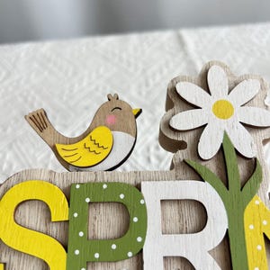 Puede incluir: Decoraci&oacute;n de madera con tem&aacute;tica primaveral que presenta un peque&ntilde;o p&aacute;jaro con detalles amarillos y marrones, una margarita blanca con centro amarillo y la palabra "SPRING" en amarillo, verde y blanco. El fondo es de veta de madera clara.