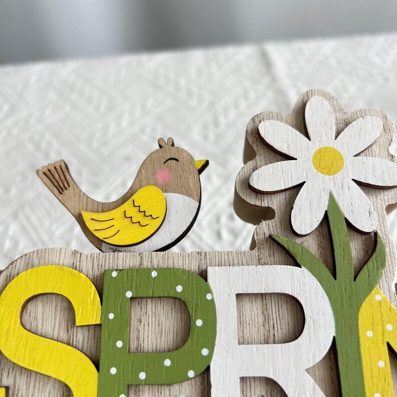 Puede incluir: Decoraci&oacute;n de madera con tem&aacute;tica primaveral que presenta un peque&ntilde;o p&aacute;jaro con detalles amarillos y marrones, una margarita blanca con centro amarillo y la palabra "SPRING" en amarillo, verde y blanco. El fondo es de veta de madera clara.