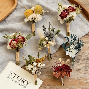 Op de afbeelding: Een assortiment kleine, gedroogde bloemboeketten. Elk boeket heeft een unieke combinatie van kleuren en texturen, waaronder rode rozen, gele pompons en witte katoen. De stelen zijn omwikkeld met touw. Een kaart met de tekst "STORY SMALL TOWN STORY" is zichtbaar.