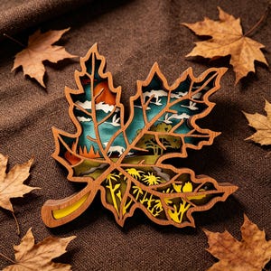 Peut inclure: Décoration en forme de feuille d'érable en bois multicouche avec une scène de paysage détaillée à l'intérieur. La feuille présente une finition en grain de bois naturel avec des accents jaunes, verts, bleus et oranges. Des feuilles séchées entourent la décoration.
