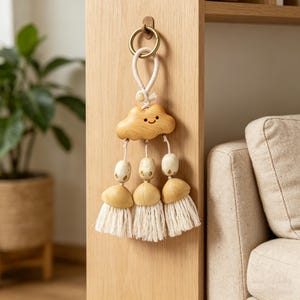 May include: A wooden cloud-shaped wall hanging with a smiling face, hanging from a brass ring. Below the cloud are three tassels with wooden beads and cream-colored fringe. The wall hanging is attached to a wooden surface.