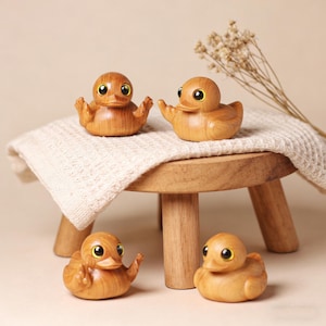 Könnte beinhalten: Vier Holz-Entenfiguren mit gelben Augen und ausgebreiteten Flügeln sind auf einem kleinen Holzschemel mit einem cremefarbenen Tuch angeordnet. Die Enten sind aus hellem Holz geschnitzt und zeigen die natürliche Maserung.