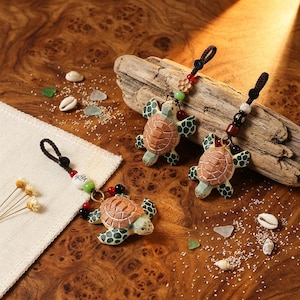 Pode incluir: Três chaveiros decorativos de tartaruga com detalhes em marrom, verde e branco. Cada tartaruga tem uma carapaça texturizada e detalhes de contas. Os chaveiros são exibidos em uma superfície de madeira com conchas e vidro do mar.