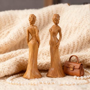 May include: Two wooden sculptures of women in elegant gowns, with intricate hairstyles. A miniature brown leather handbag and a pearl necklace are also visible. The sculptures are displayed against a cream-coloured, textured backdrop.