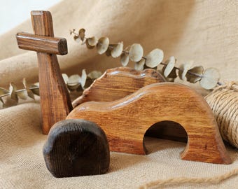 May include: Wooden religious scene featuring a cross, a curved arch, a small hill, and a dark brown stone. The items are made of wood with varying shades of brown and are arranged on a tan fabric surface.