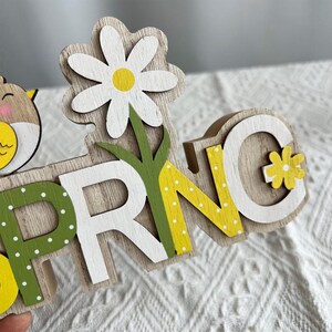 Puede incluir: Letrero de madera con la palabra "SPRING" en blanco, verde y amarillo. El letrero "SPRING" tiene una margarita y un peque&ntilde;o b&uacute;ho. Las letras est&aacute;n pintadas con lunares y el letrero es de madera.