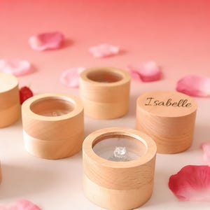 May include: Wooden ring boxes with clear tops, one containing a diamond ring. The boxes are cylindrical with a light wood finish and a contrasting base. One box is engraved with the name "Isabelle". Pink rose petals are scattered around the boxes.