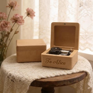 May include: A wooden music box with the lid open, revealing the mechanical components. The box is engraved with the words "To Alice". Another closed wooden box is next to it. The boxes are on a table with a white lace cloth.