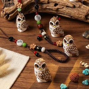 Pode incluir: Uma coleção de estatuetas de corujas e acessórios com contas. As corujas são brancas e marrons com marcações detalhadas. Os acessórios incluem cordões e pulseiras de contas com contas e berloques decorativos. Os itens estão dispostos em uma superfície de madeira.