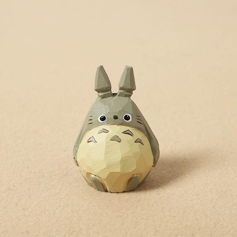 May include: A wooden figurine of Totoro, a character from the animated film "My Neighbor Totoro." The figure is gray and tan, with large eyes and pointed ears. The figurine is carved with visible wood grain.