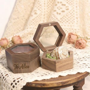 May include: Hexagonal wooden ring box with a clear top, open to reveal a large, ornate ring. The box is engraved with "To Alice." The ring is set on a bed of green moss. Soft pink roses are in the background.