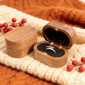 Könnte beinhalten: Holzringbox mit einem Silberring darin, auf einer cremefarbenen Strickoberfläche. Die Box ist mit den Worten "To Alice" graviert. Ein Zweig mit roten Beeren befindet sich im Hintergrund.