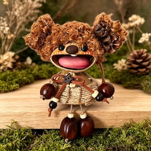 May include: A handcrafted doll with a distinctive appearance. The doll has brown, curly hair-like features, a smiling face with a pink tongue, and decorative accents. The doll's body is made of woven material, and it has acorn-like feet.