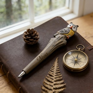 Peut inclure: Un stylo en bois avec une figurine de hibou, le mot "Nova" est gravé sur le stylo. Une petite boussole, une pomme de pin et une fougère sont sur un journal en cuir marron. Le stylo est un accessoire de bureau unique.