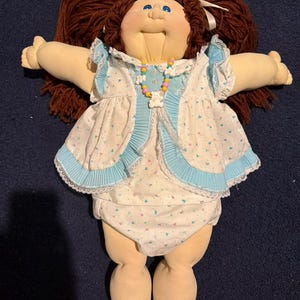 Könnte beinhalten: Eine Vintage-Puppe mit braunen Wollhaaren, die zu Zöpfen geflochten sind. Die Puppe trägt ein weißes Kleid mit blauer Bordüre und eine passende Windel. Eine bunte Perlenkette schmückt den Hals der Puppe. Die Arme der Puppe sind ausgestreckt.