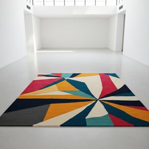 May include: A rectangular area rug with a geometric design. The rug features a vibrant pattern of triangles and shapes in shades of red, yellow, blue, and white. The rug is placed on a light-colored floor in a spacious, well-lit room.