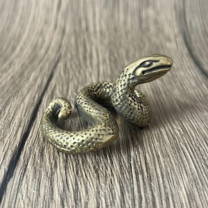 Może przedstawiać: Brązowa figurka węża ze szczegółowymi łuskami i zwiniętym ciałem. Głowa węża jest uniesiona, a jego oczy są widoczne. Figurka jest wykonana z metalu i ma teksturowaną powierzchnię.