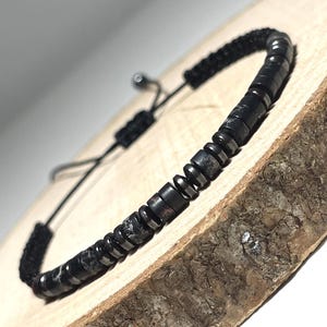 Peut inclure: Bracelet de perles noires avec fermeture coulissante. Le bracelet est composé de petites perles rondes et sombres et d'un cordon noir. Les perles ont une finition mate et sont enfilées ensemble. Le bracelet est présenté sur une surface en bois.