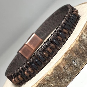 May include: A brown leather bracelet with a magnetic clasp. The bracelet has a woven design with alternating black and copper-coloured beads. The clasp is rectangular with a copper finish. The bracelet is displayed on a wooden surface.