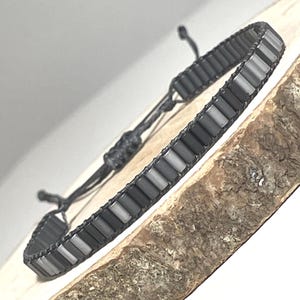 Könnte beinhalten: Ein dunkelgraues Perlenarmband mit verstellbaren schwarzen Kordelbändern. Das Armband hat rechteckige Perlen in verschiedenen Grautönen, die zu einem flexiblen Band aufgereiht sind. Das Armband wird auf einer Holzoberfläche präsentiert.