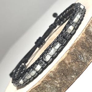 May include: A wrap bracelet featuring a combination of black and gray square beads and white rectangular stones. The bracelet is strung on a black cord with an adjustable knot closure. The bracelet is displayed on a wooden surface.