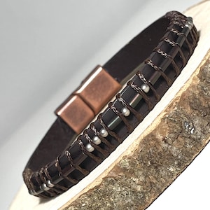 May include: A dark brown leather bracelet with a magnetic clasp. The bracelet features dark rectangular beads woven with brown cord and accented with small silver beads. The clasp is a copper colour.