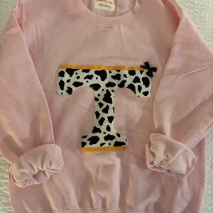 Puede incluir: Una sudadera rosa claro con una gran letra "T" aplicada en un estampado de vaca en blanco y negro. La parte superior de la "T" está bordeada con tela amarilla. Un pequeño lazo negro está unido al lado derecho de la "T". La etiqueta dice "Loved By Lynn Stitchery".