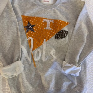 May include: A gray sweatshirt with rolled-up sleeves. The front features an orange pennant with white polka dots, a star, and a football-shaped patch. The word "Vols" is appliquéd in white, and the letter "T" is visible.