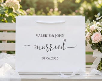 May include: A white rectangular gift bag with white cord handles featuring the printed text VALERIE & JOHN, the word married in a cursive script, and the date 07.06.2026 in a dark grey font, positioned next to a bouquet of light pink roses.
