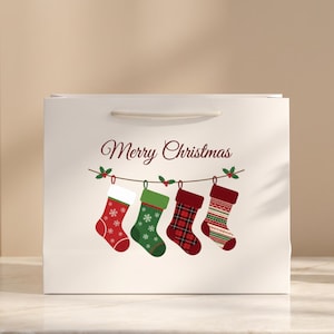 Può includere: Una busta regalo rettangolare bianca con la scritta Merry Christmas in corsivo rosso sopra un disegno di quattro calze natalizie appese. Le calze includono una rossa con fiocchi bianchi, una verde con fiocchi, una a quadri rossi e una a righe.