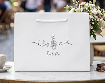 Op de afbeelding: Een witte rechthoekige cadeauzak met witte koordhengsels. De voorzijde is voorzien van een zwarte lijntekening van een vioolsleutel en muzieknoten, met de naam Isabelle in een cursief lettertype onder de muzikale afbeelding gedrukt.