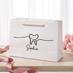 May include: A white gift bag featuring a black continuous line drawing of a tooth and the name Sophia printed in black script. The bag includes a light pink ribbon handle and is accompanied by a matching pink satin ribbon curled nearby.