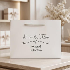 May include: A white rectangular gift bag with a top handle featuring the black printed text Liam & Chloe, a small heart graphic, the word engaged, and the date 02.06.2026 centred on the front surface of the smooth matte paper material.