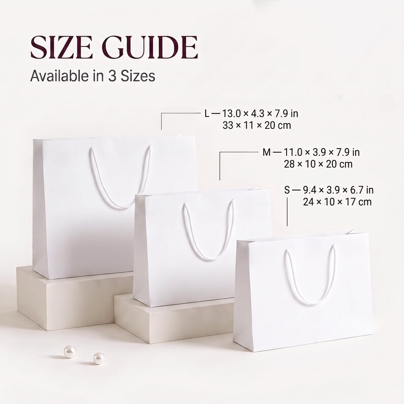 May include: Three plain white paper gift bags with white cord handles, displayed in small, medium, and large sizes. Text reads SIZE GUIDE Available in 3 Sizes. Dimensions are listed as L 33 x 11 x 20 cm, M 28 x 10 x 20 cm, and S 24 x 10 x 17 cm.