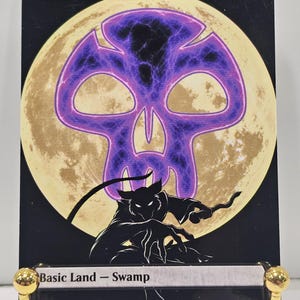 Op de afbeelding: Een rechthoekige ruilkaart met de titel Swamp, met een groot, gloeiend paars schedelpictogram gecentreerd over een lichtgele volle maan. Onder de maan staat een zwart silhouet van een gebogen, klauwend wezen tegen een donkere achtergrond. De tekst luidt Swamp en Basic Land - Swamp.
